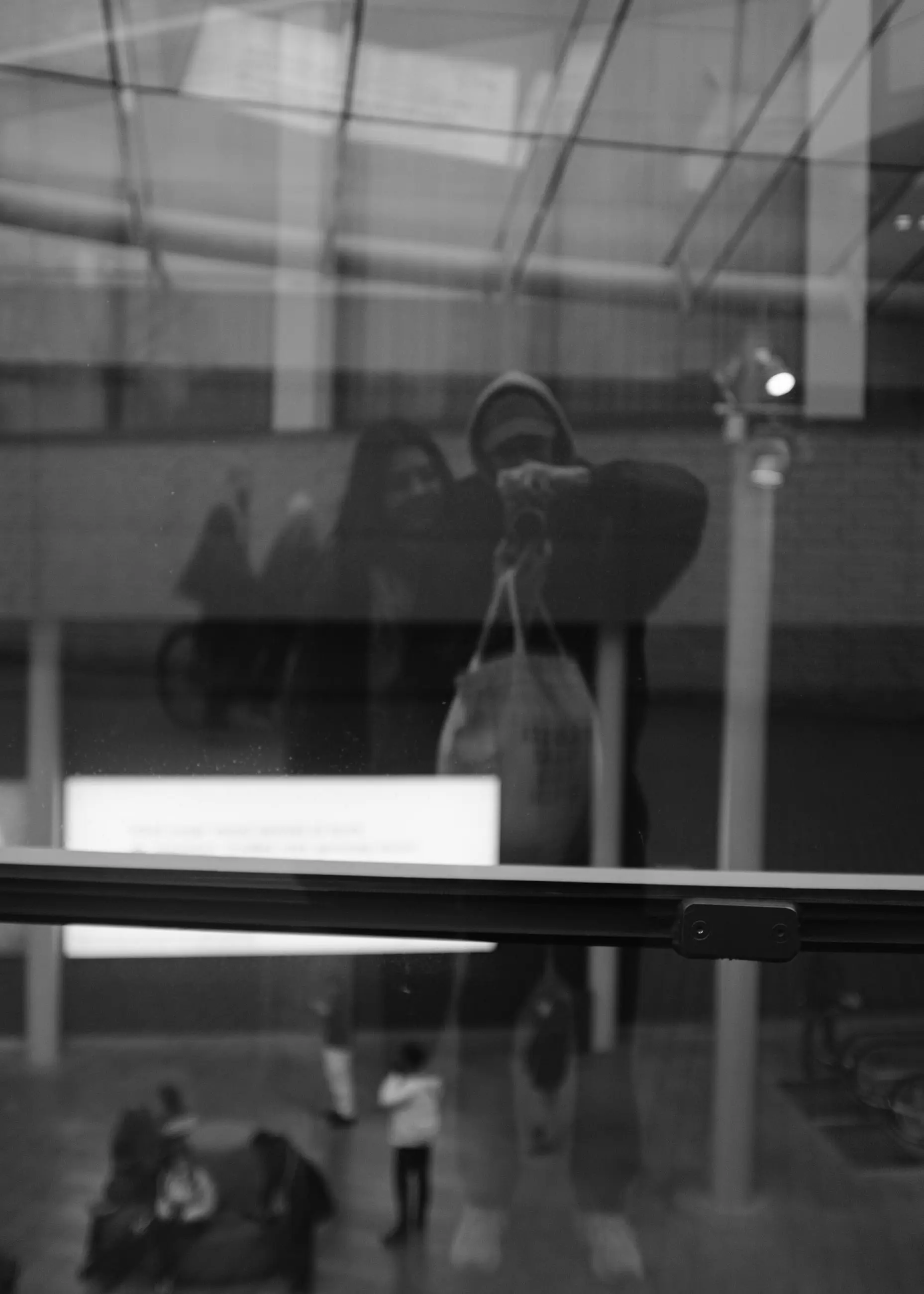 A figure reflected in dark museum glass, visitors on a bench beyond