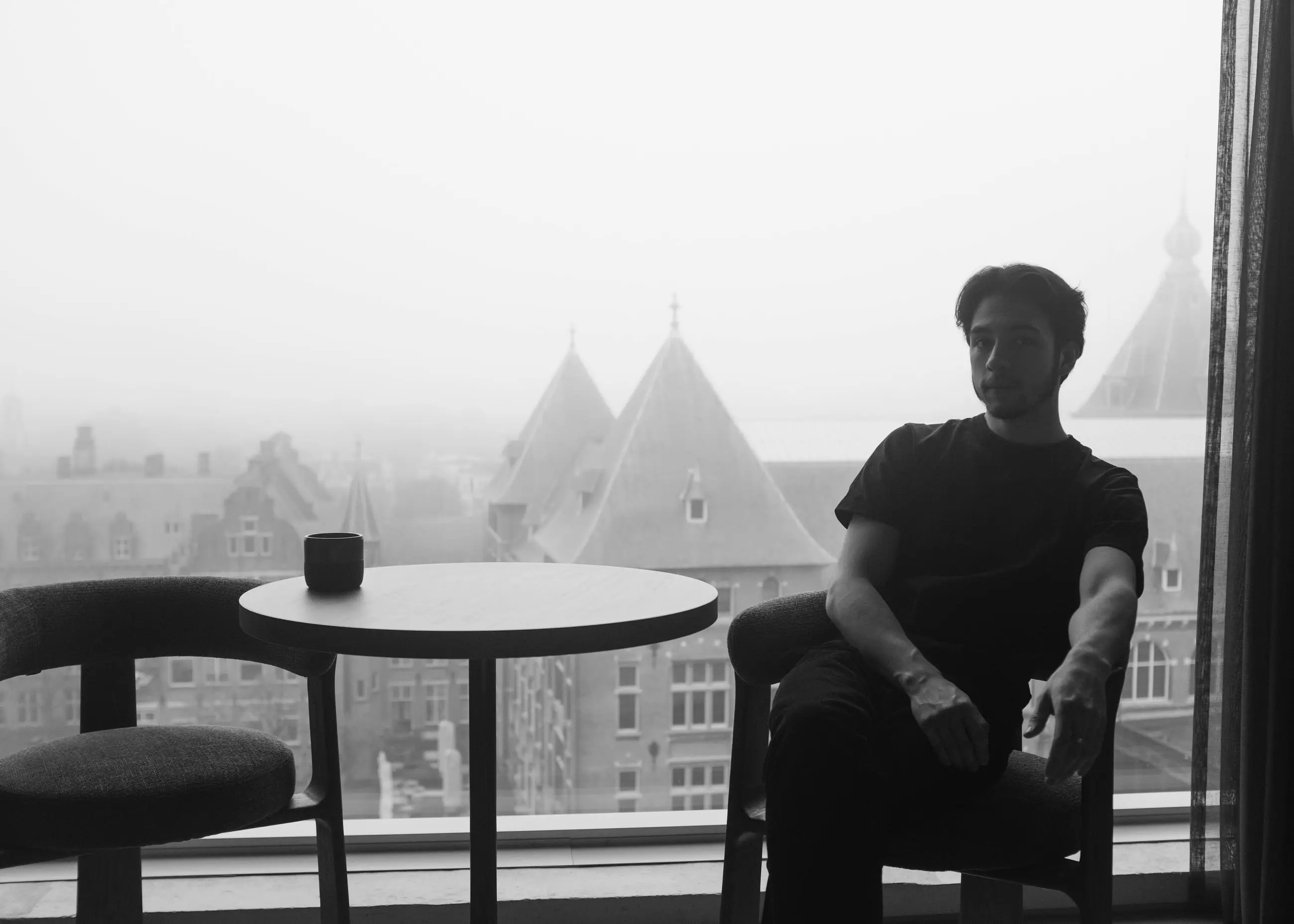A man seated at the window, a cup on the table, fog over the roofs