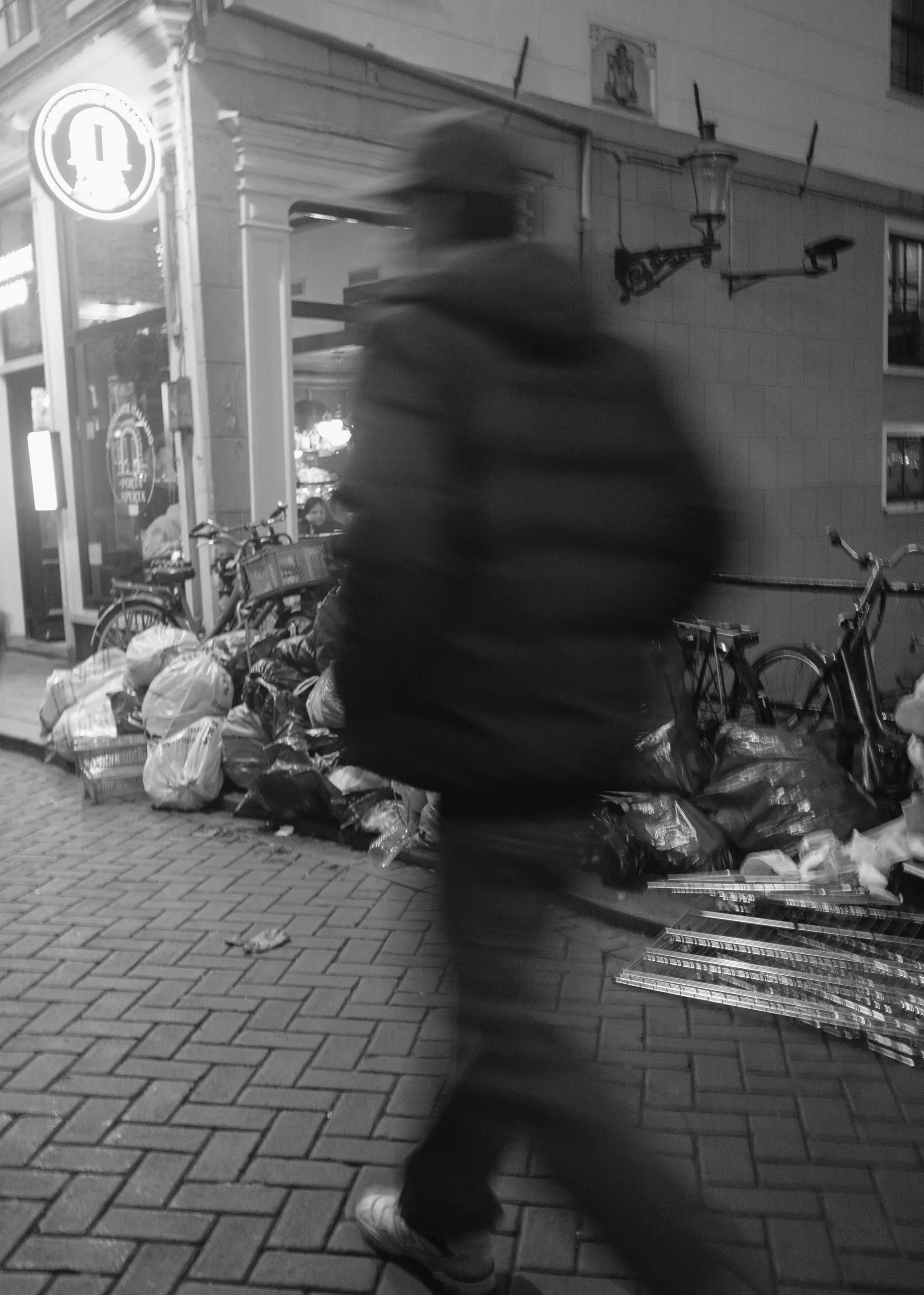 A walker blurred past a lit doorway and rubbish bags